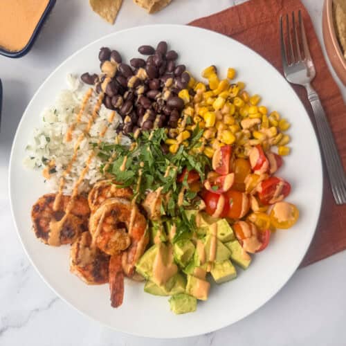 Overhead view of white plate with a shrimp burrito bowl made with seasoned shrimp, cilantro rice, black beans, diced avocado, cherry tomatoes, and corn garnished with cilantro and chipotle almond drizzle. Additional chipotle almond dressing, tortilla chips, a napkin and a fork surround the plate.