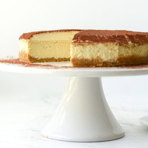 Side view of a cut Tiramisu Cheesecake on a cake stand next to a shaker of cocoa powder