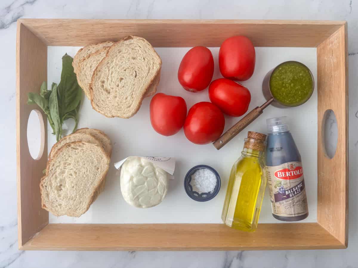 Overhead photo of individual ingredients needed to make caprese bruschetta: sourdough bread, Roma tomatoes, mozzarella pearls, fresh basil, pesto, olive oil, balsamic glaze and salt. 