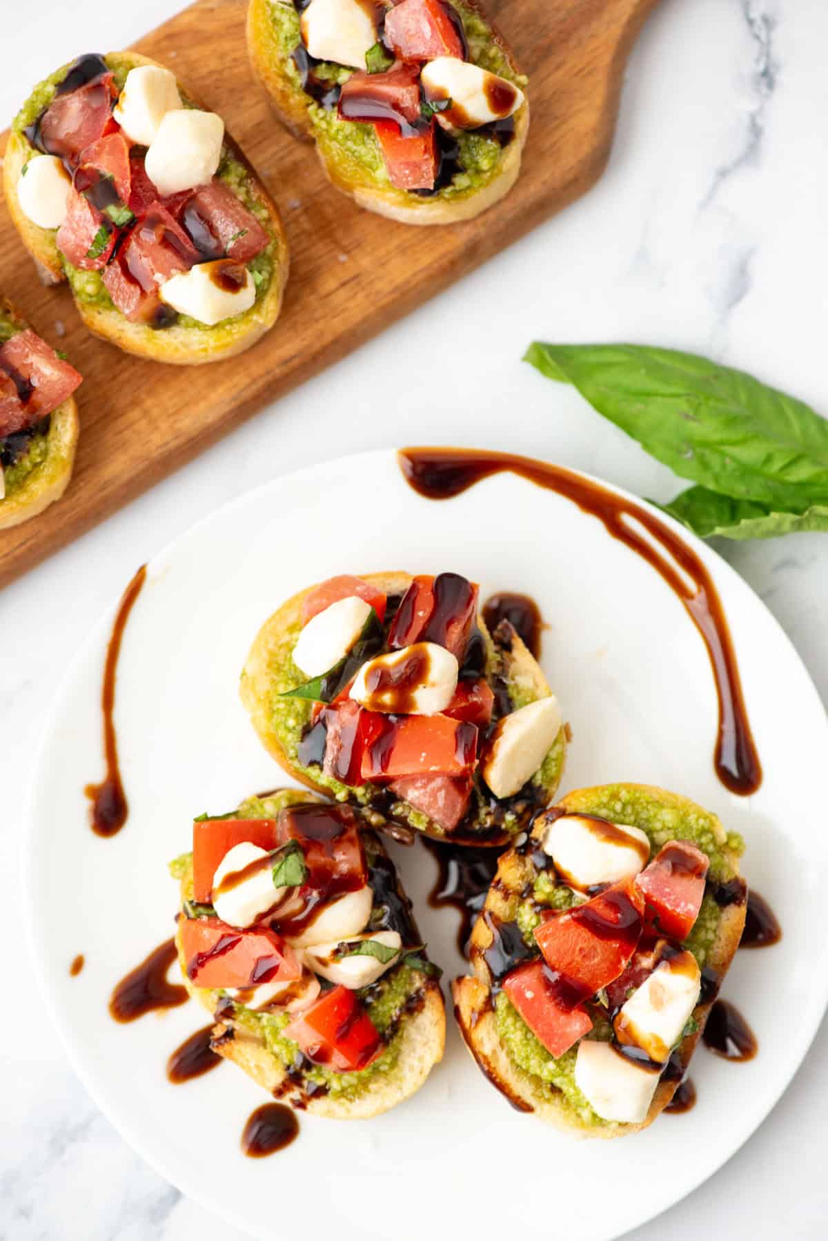 Three pieces of caprese baguette on a plate with balsamic drizzle in front of a serving board with additional caprese bruschetta