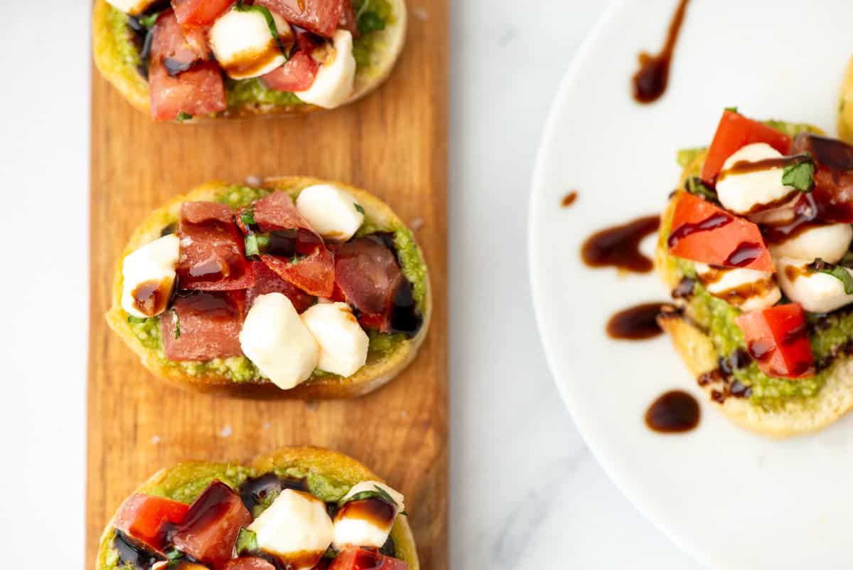 Pieces of caprese bruschetta on a board next to a plate with additional pieces of caprese bruschetta. All drizzled with balsamic glaze.