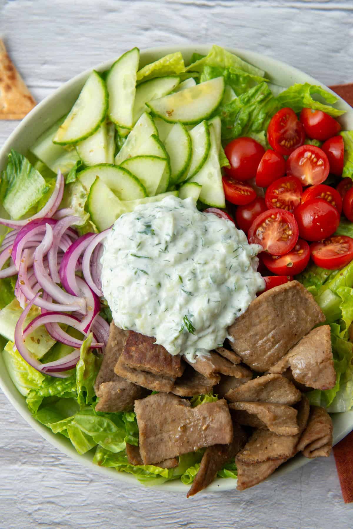 Overhead view of gyro salad made with romaine lettuce, gyro meat, cherry tomatoes, cucumbers, red onion, a light Greek dressing and a helping of tzatziki sauce on a large salad plate