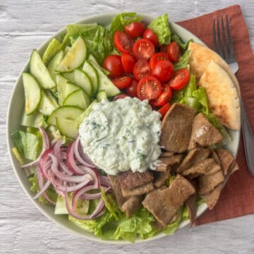 Overhead view of gyro salad made with romaine lettuce, gyro meat, cherry tomatoes, cucumbers, red onion, a light Greek dressing and a helping of tzatziki sauce on a plate next to a napkin and fork