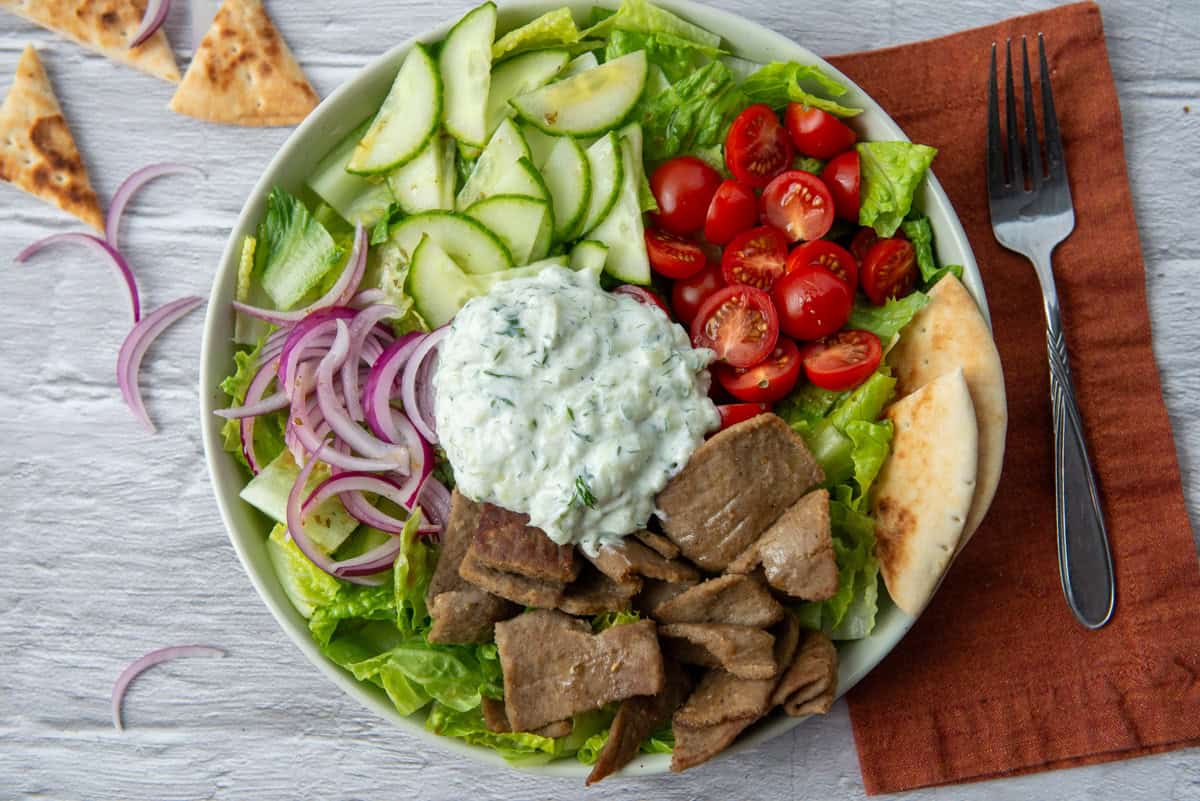 Overhead view of gyro salad made with romaine lettuce, gyro meat, cherry tomatoes, cucumbers, red onion, a light Greek dressing and a helping of tzatziki sauce on a plate next to a napkin, fork and scatter pita chips and red onion 
