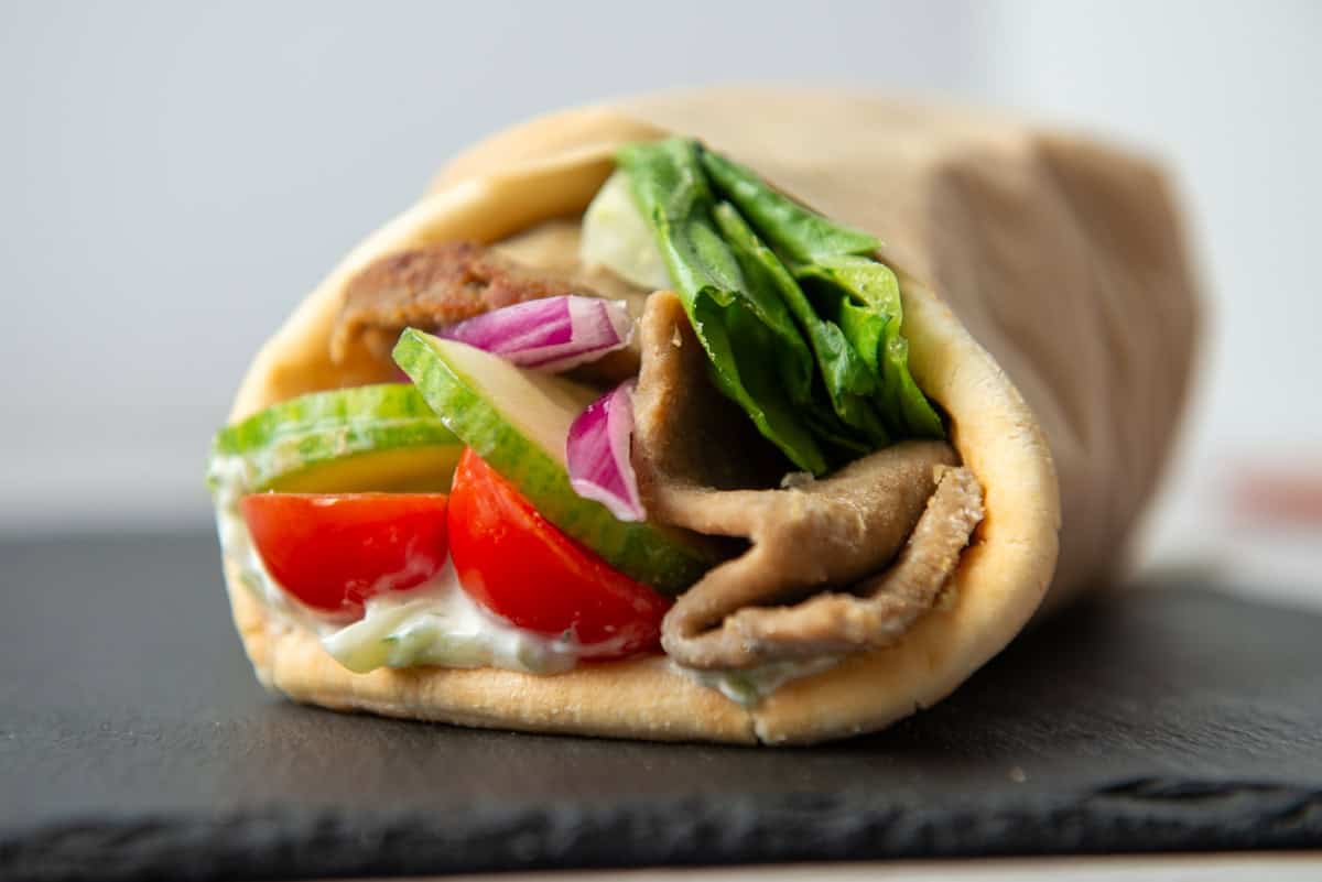Side view of a gyro sandwich containing tzatziki, tomatoes, lettuce cucumbers and onions wrapped in parchment paper on a slate board
