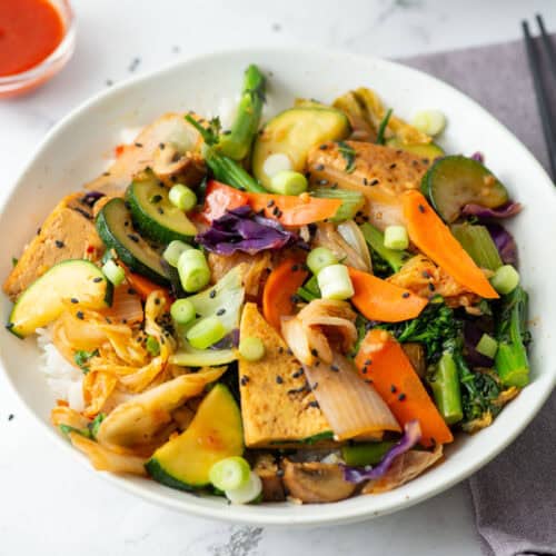 Overhead view of a bowl of kimchi tofu stir fry with vegetables over rice with chopsticks on the side and a tiny bowl of sriracha in the background