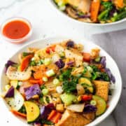 Overhead view of a bowl of kimchi tofu stir fry with vegetables over rice with a tiny bowl of sriracha and another bowl of kimchi tofu stir fry in the background