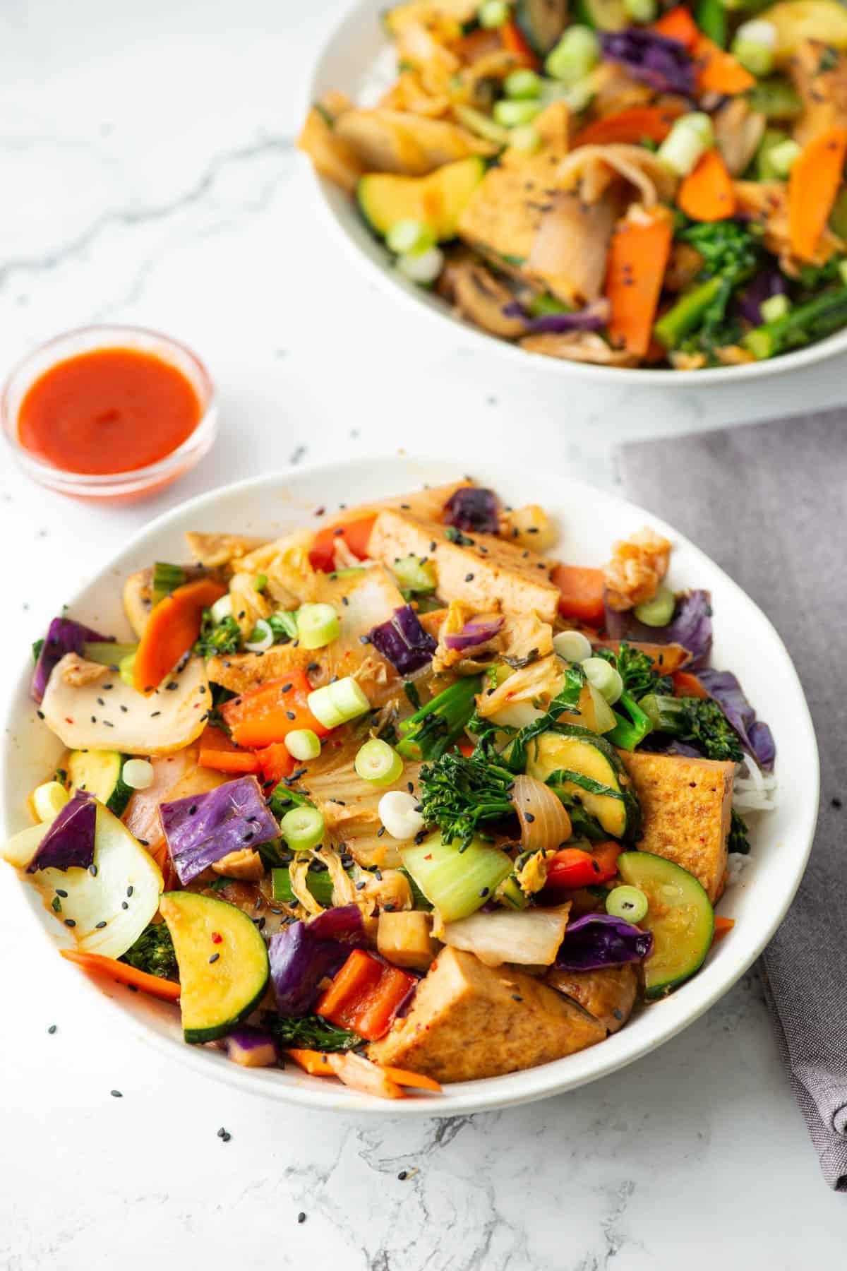 Overhead view of a bowl of kimchi tofu stir fry with vegetables over rice with a tiny bowl of sriracha and another bowl of kimchi tofu stir fry in the background