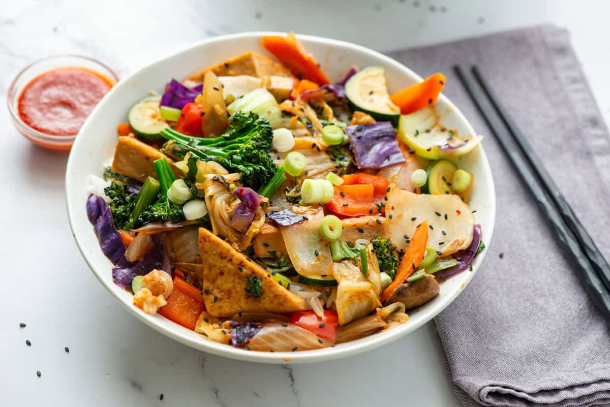 Overhead view of a bowl of kimchi tofu stir fry with vegetables over rice with a napkin, chopsticks and a tiny bowl of sriracha on the side
