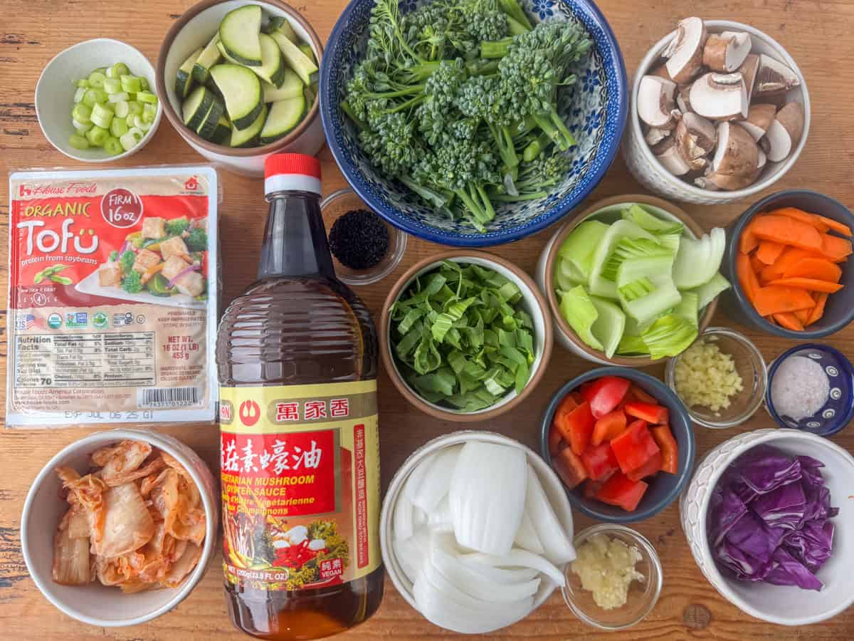 Ingredients needed to make kimchi tofu stir fry: kimchi, tofu, onions, mushrooms, bok choy, red peppers, carrots, red cabbage, zucchini, broccolini, stir fry sauce, garlic, ginger and salt. Also shown are optional garnishes of sliced green onions and black sesame seeds.
