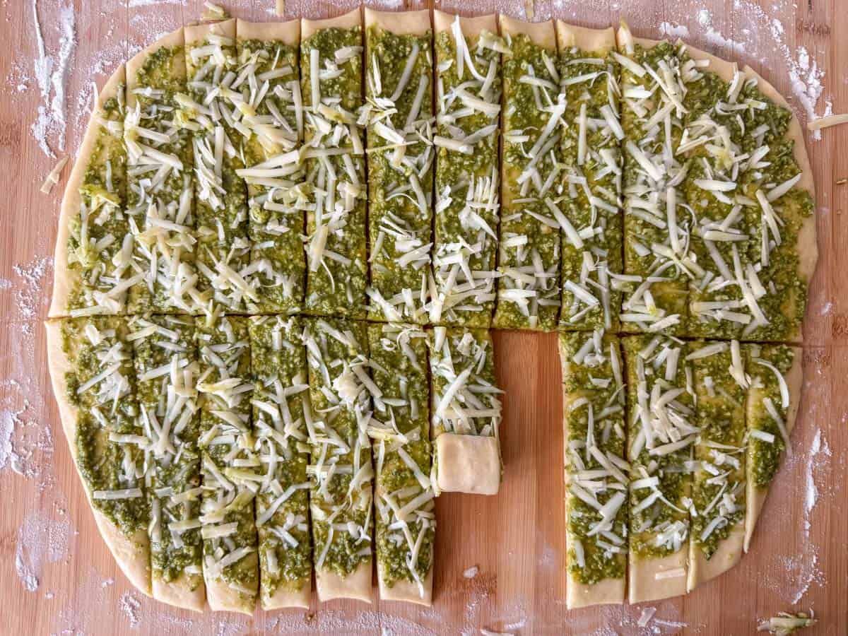 Overhead view of rolled out uncooked dough, topped with pesto and gruyere cheese and then cut into strips with a pizza cutter