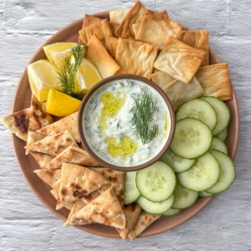 Overhead view of a small bowl of tzatziki dip in the serving of a serving tray that contains pita chips, cucumbers, pita triangles and lemon wedges