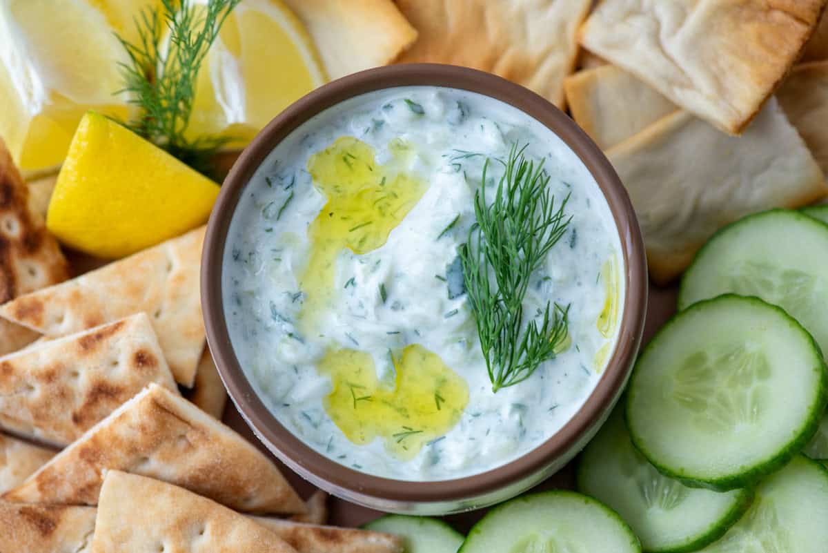 Overhead view of a small bowl of tzatziki dip in the serving of a serving tray that contains pita chips, cucumbers, pita triangles and lemon wedges