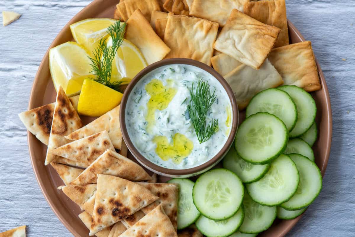 Overhead view of a small bowl of tzatziki dip in the serving of a serving tray that contains pita chips, cucumbers, pita triangles and lemon wedges
