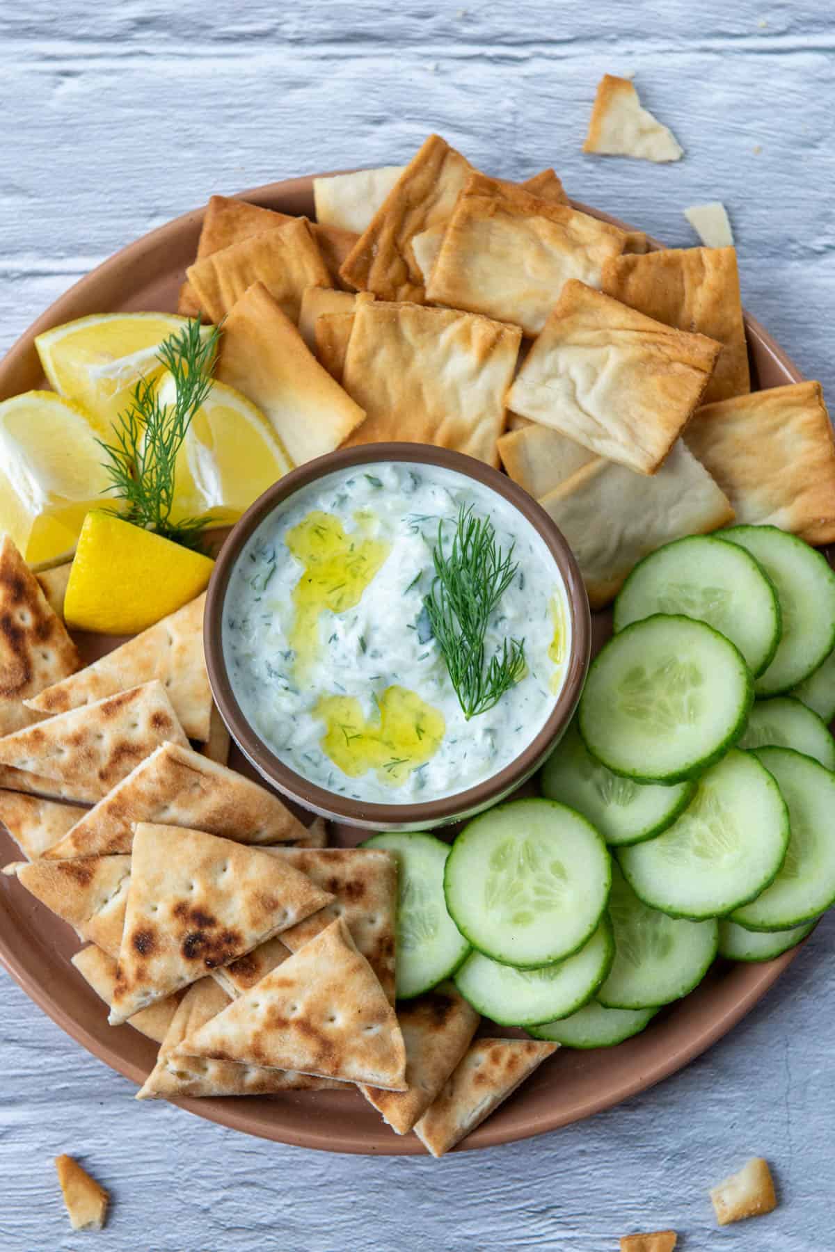 Overhead view of a small bowl of tzatziki dip in the serving of a serving tray that contains pita chips, cucumbers, pita triangles and lemon wedges