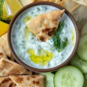 Overhead view of a small bowl of tzatziki dip with a pita triangle sticking out surrounded by pita chips, cucumbers, pita triangles and lemon wedges