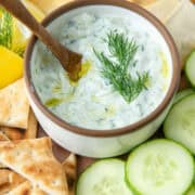 Overhead view of a small bowl of tzatziki dip surrounded by pita chips, cucumbers, pita triangles and some lemon wedges