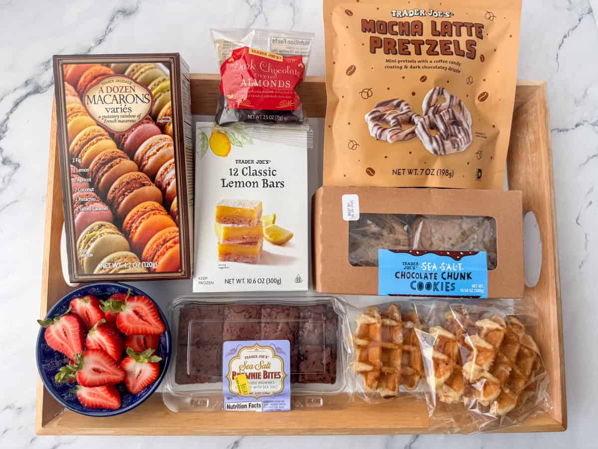 Overhead view of the ingredients needed to make a dessert board: brownie bites, lemon bars, chocolate chip cookies, macarons, mocha latte pretzels, Belgian waffles, chocolate covered almonds and strawberries.