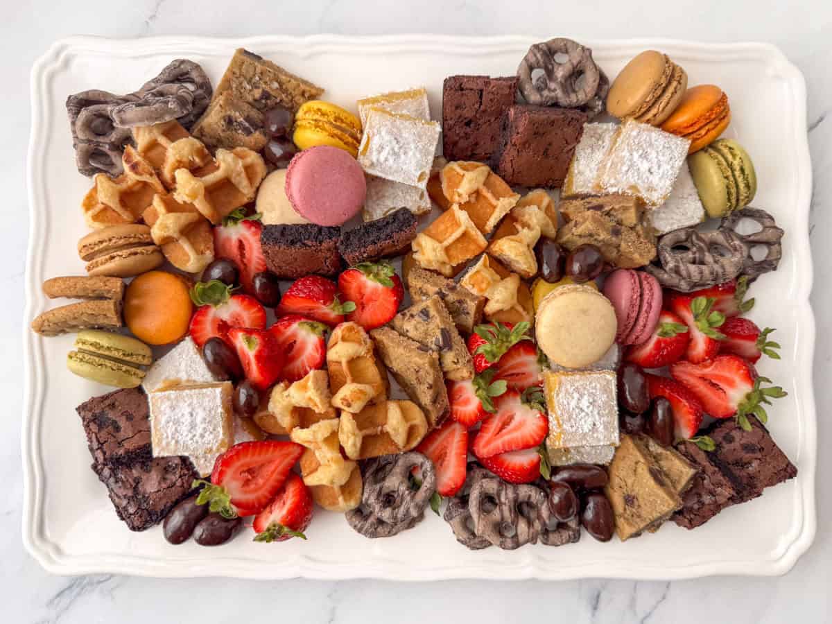 Overhead view of a dessert board made with brownie bites, lemon bars, chocolate chip cookies, macarons, mocha latte pretzels, Belgian waffles, chocolate covered almonds and strawberries.  