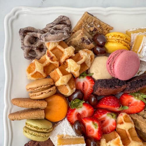Close up photo of a corner of a dessert board made with brownie bites, lemon bars, chocolate chip cookies, macarons, mocha latte pretzels, Belgian waffles, chocolate covered almonds and strawberries