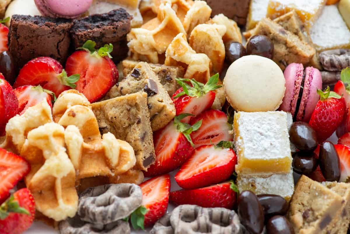 Close up photo of a dessert board made with brownie bites, lemon bars, chocolate chip cookies, macarons, mocha latte pretzels, Belgian waffles, chocolate covered almonds and strawberries