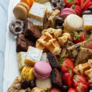 Close up photo of a corner of a dessert board made with brownie bites, lemon bars, chocolate chip cookies, macarons, mocha latte pretzels, Belgian waffles, chocolate covered almonds and strawberries