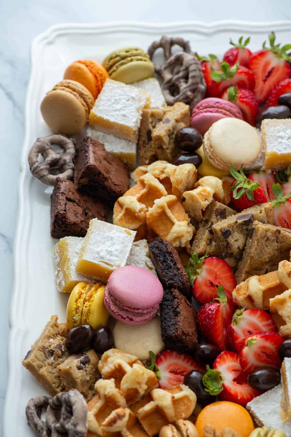 Close up photo of a corner of a dessert board made with brownie bites, lemon bars, chocolate chip cookies, macarons, mocha latte pretzels, Belgian waffles, chocolate covered almonds and strawberries