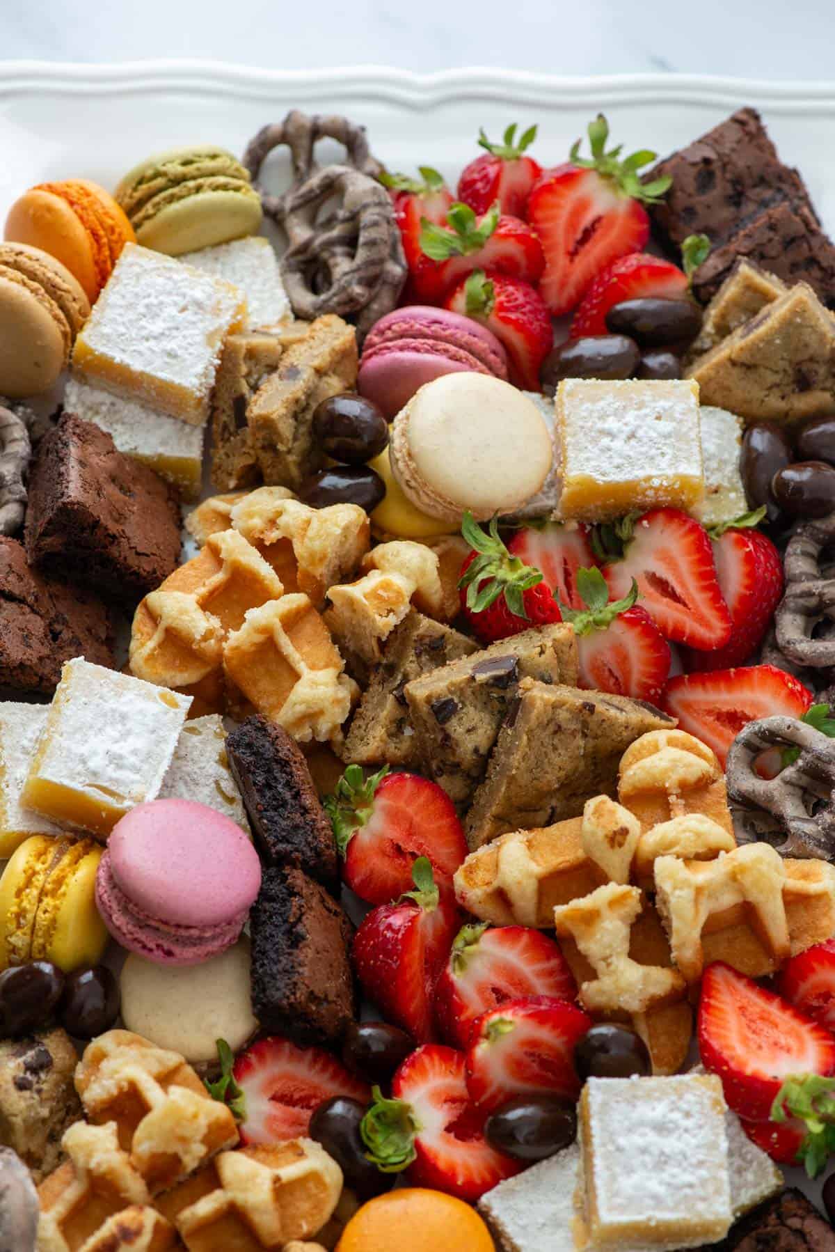 Close up photo of a dessert board made with brownie bites, lemon bars, chocolate chip cookies, macarons, mocha latte pretzels, Belgian waffles, chocolate covered almonds and strawberries