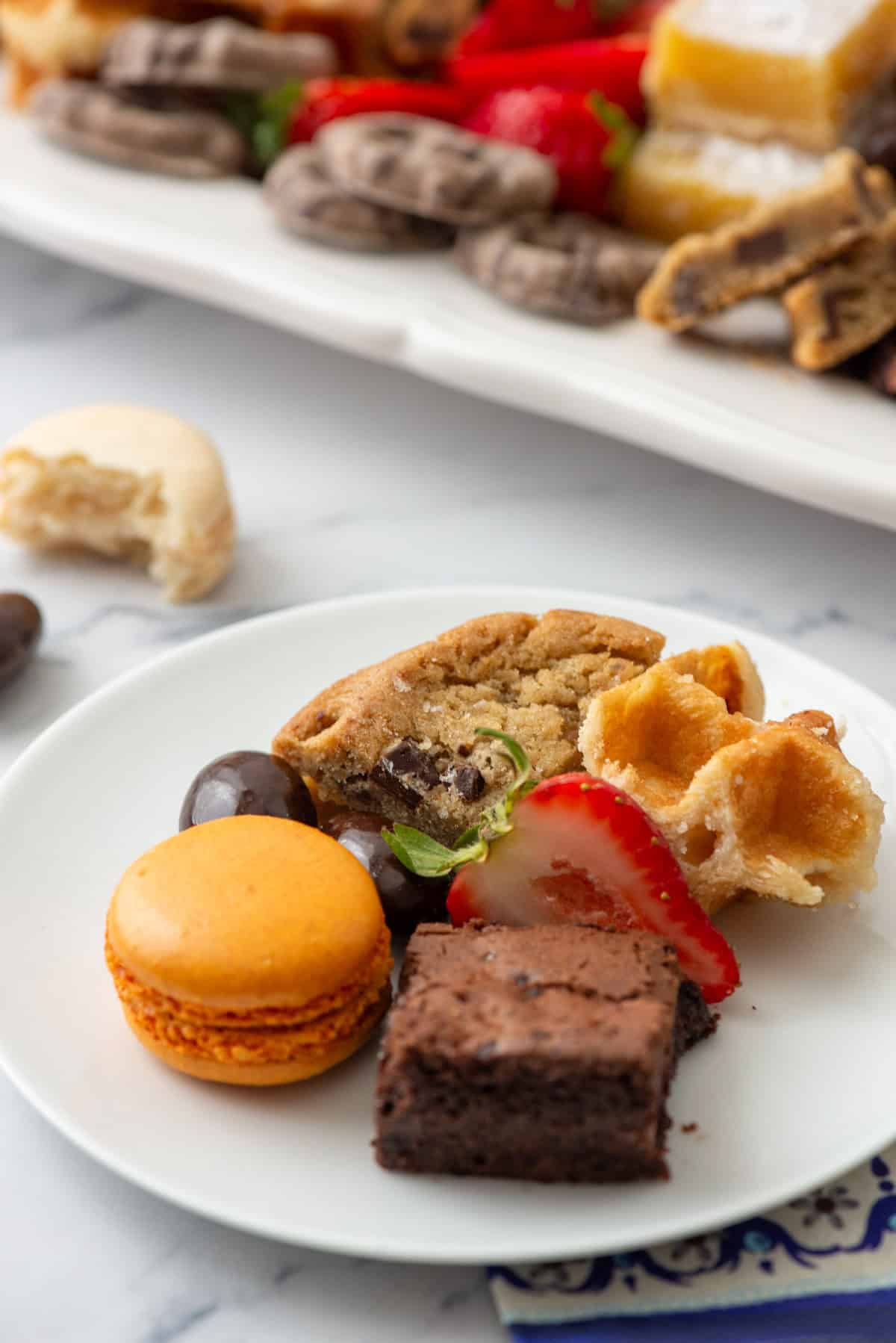 Close up photo of a small dessert plate with an orange macaron, brownie bite, cut strawberry, Belgian waffle piece, chocolate chip cookie and chocolate covered almond with a dessert board in the background