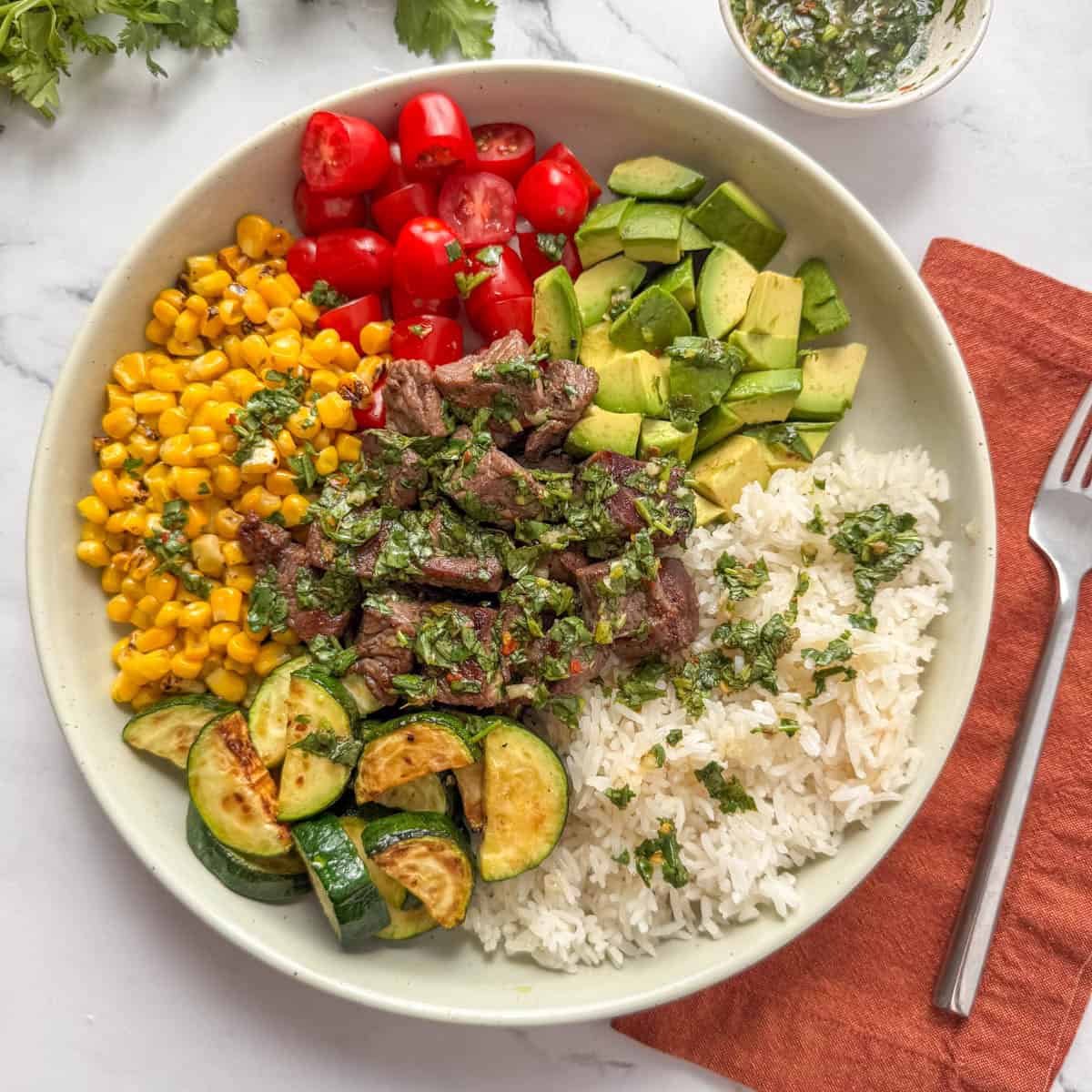 Overhead photo of a steak and rice bowl made with steak bites, rice, zucchini, corn, avocado, cherry tomatoes and chimichurri sauce.