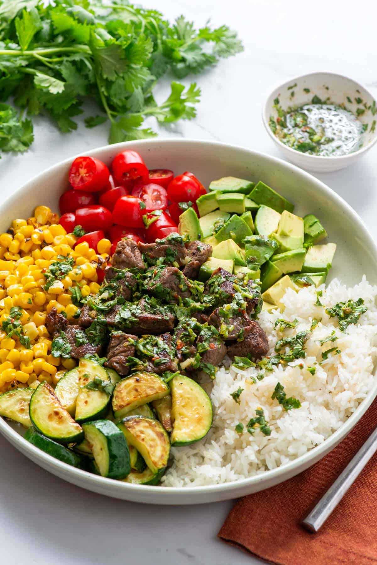 Overhead photo of a steak and rice bowl made with steak bites, rice, zucchini, corn, avocado, cherry tomatoes and chimichurri sauce.