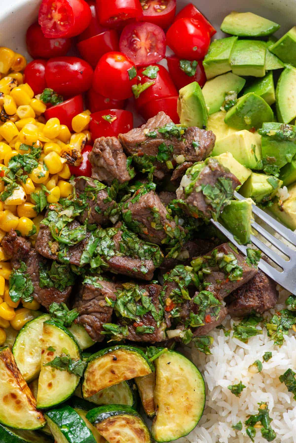 Close up view of cilantro chimichurri over steak in a rice bowl with corn, zucchini, avocado and cherry tomatoes