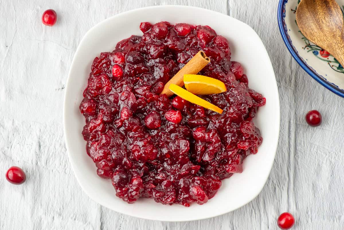Overhead view of a bowl of Grand Marnier cranberry sauce garnished with orange slices and a cinnamon stick surrounded by some scattered fresh cranberries and a spoon on a spoon rest