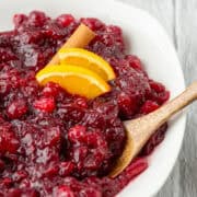 Overhead view of a partial bowl of Grand Marnier cranberry sauce garnished with orange slices and a cinnamon stick with a spoon sticking out and surrounded by some scattered fresh cranberries