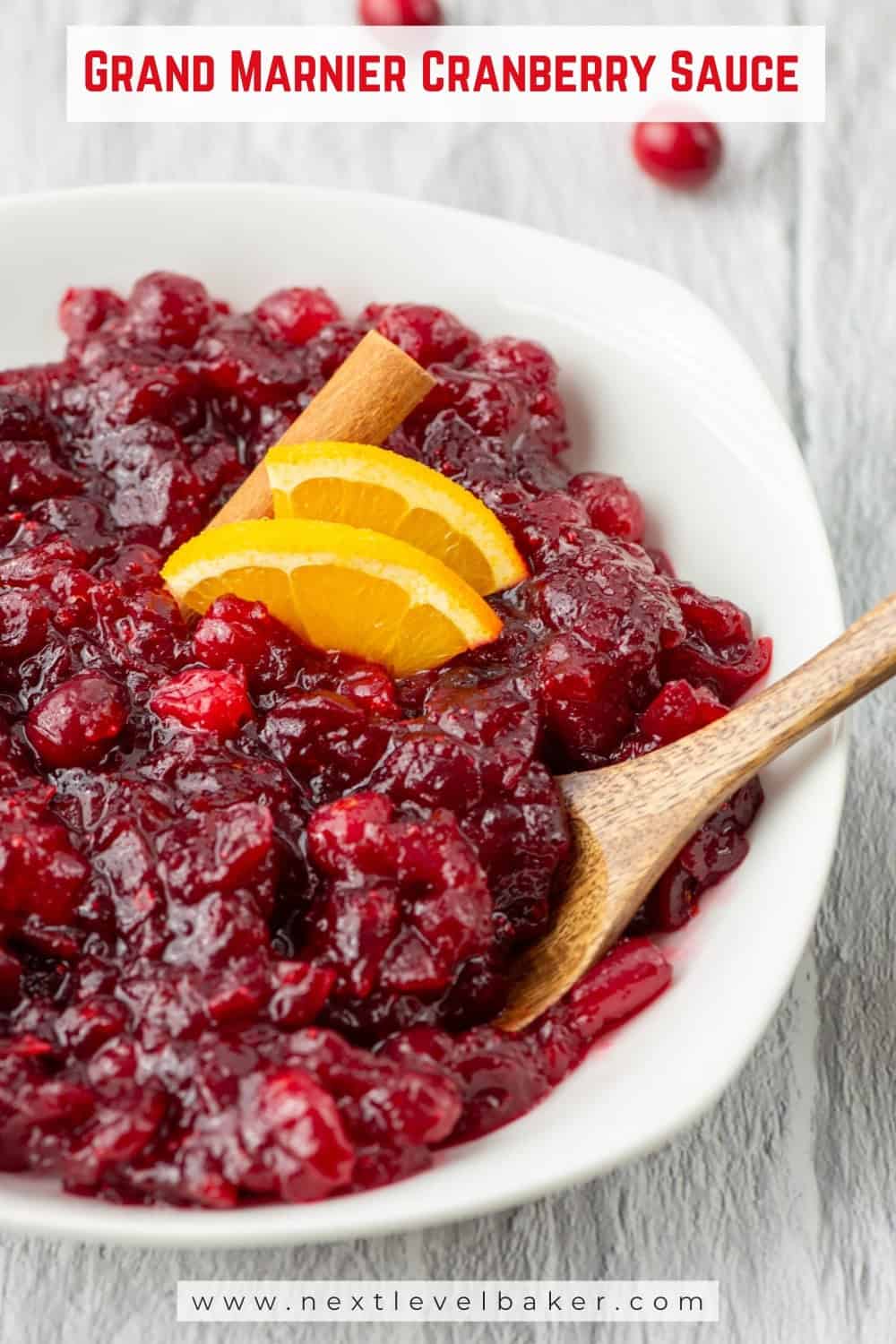 Overhead view of a partial bowl of Grand Marnier cranberry sauce garnished with orange slices and a cinnamon stick with a spoon sticking out and surrounded by some scattered fresh cranberries with a title overlay that says Grand Marnier Cranberry Sauce and www.nextlevelbaker.com