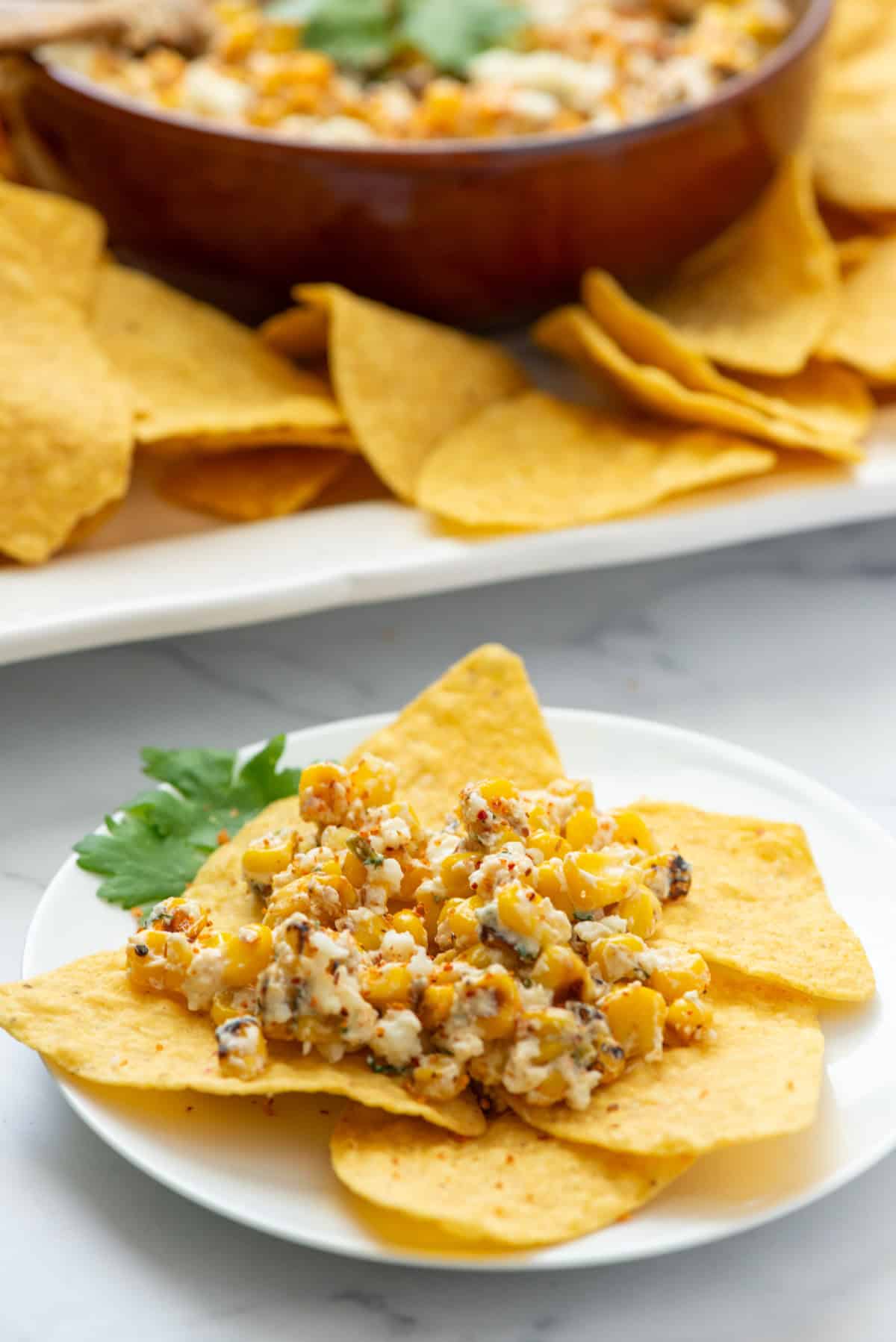 Mexican street dip on tortilla chips on a plate in front of a bowl of Mexican street corn dip