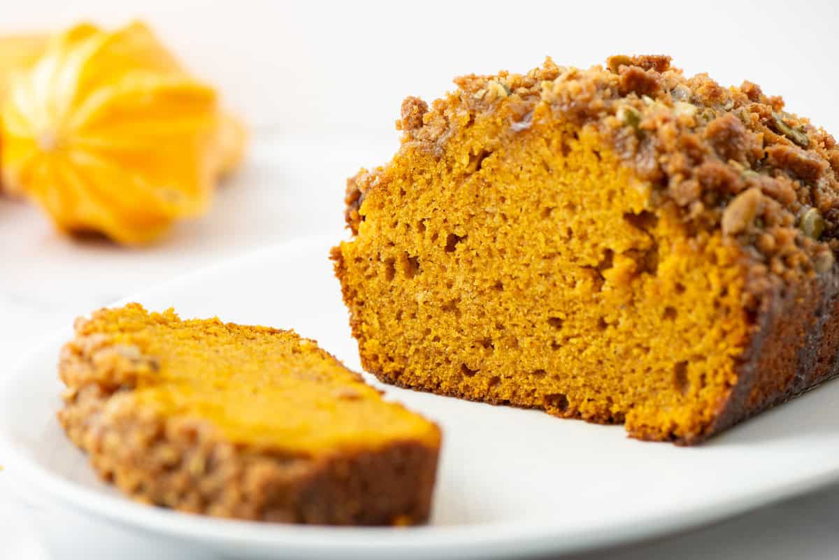 Slice of pumpkin bread on a plate in front of the pumpkin loaf with a pumpkin gourd in the background