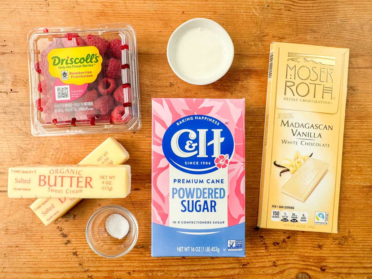 Ingredients needed to make white chocolate buttercream frosting: salted butter, white chocolate, powdered sugar, heaving whipping cream, salt and fresh raspberries for garnish.