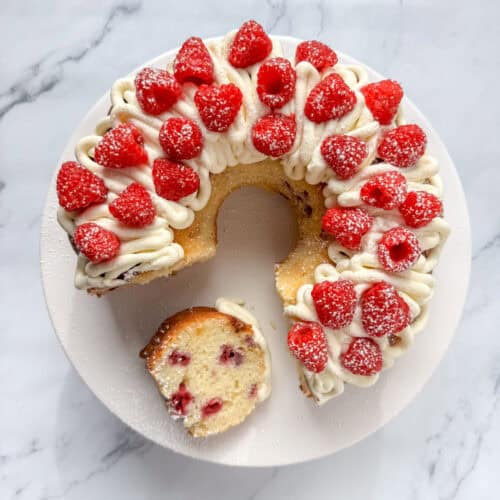 Overhead view of a white chocolate raspberry bundt cake with white chocolate buttercream frosting and fresh raspberries with a slice cut out and on its side