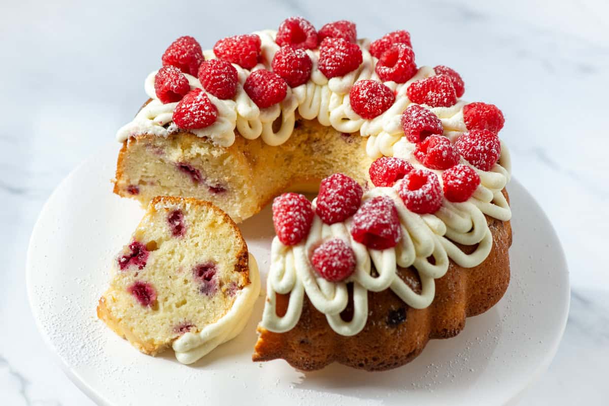 Overhead view of a white chocolate raspberry bundt cake with white chocolate buttercream frosting and fresh raspberries with a slice cut out and on its side