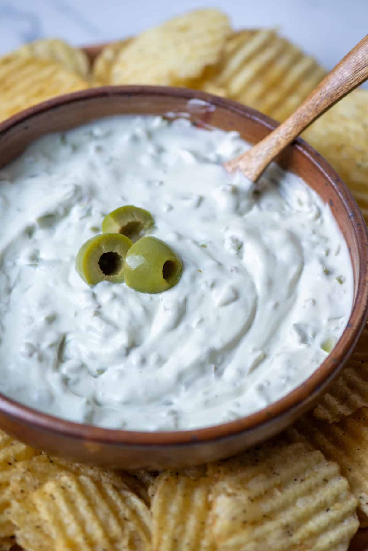 Overhead photo of dirty martini dip with a serving spoon sticking out and garnished with a few olive slices and surrounded by kettle style potato chips.