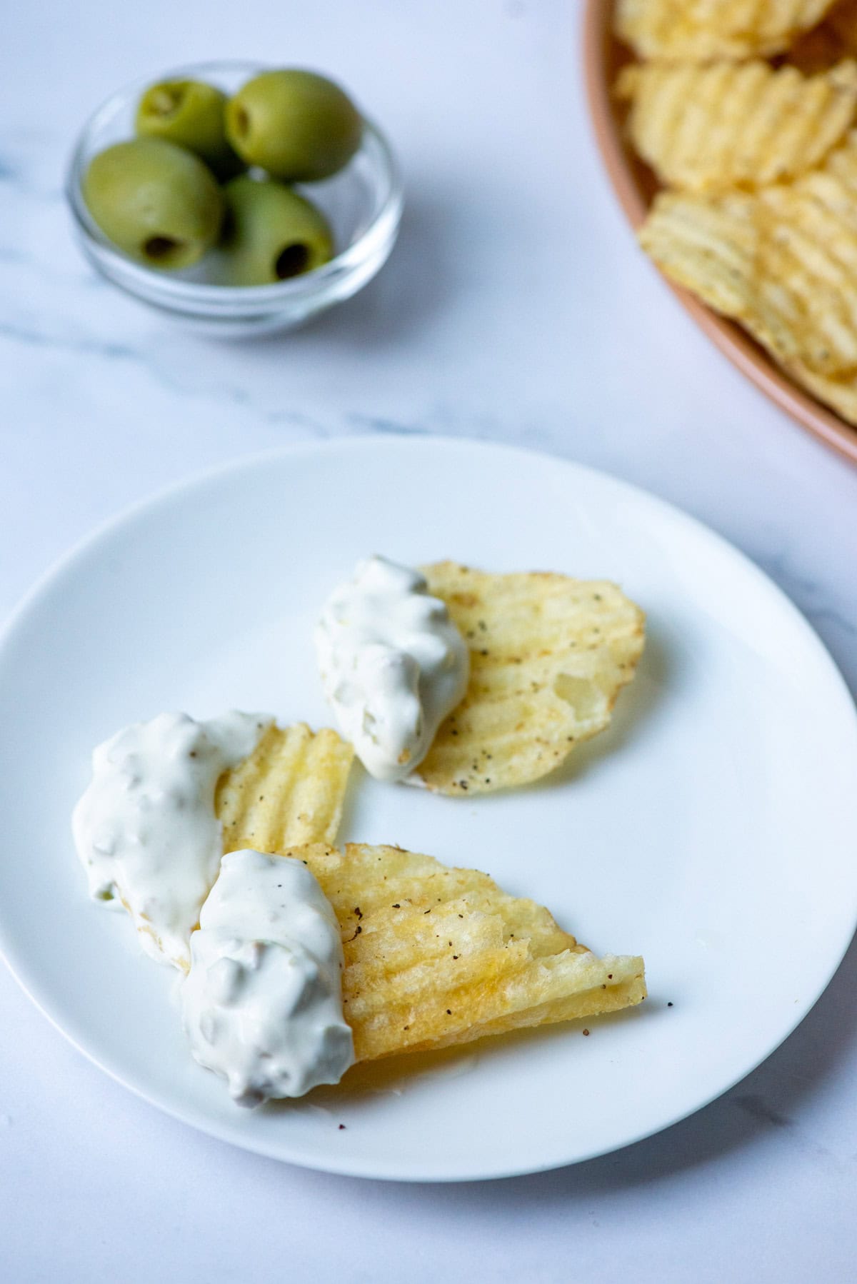 Three chips dipped in Dirty Martini Dip on a plate in front of a bowl of chips and a tiny sauce cup of olives in the background