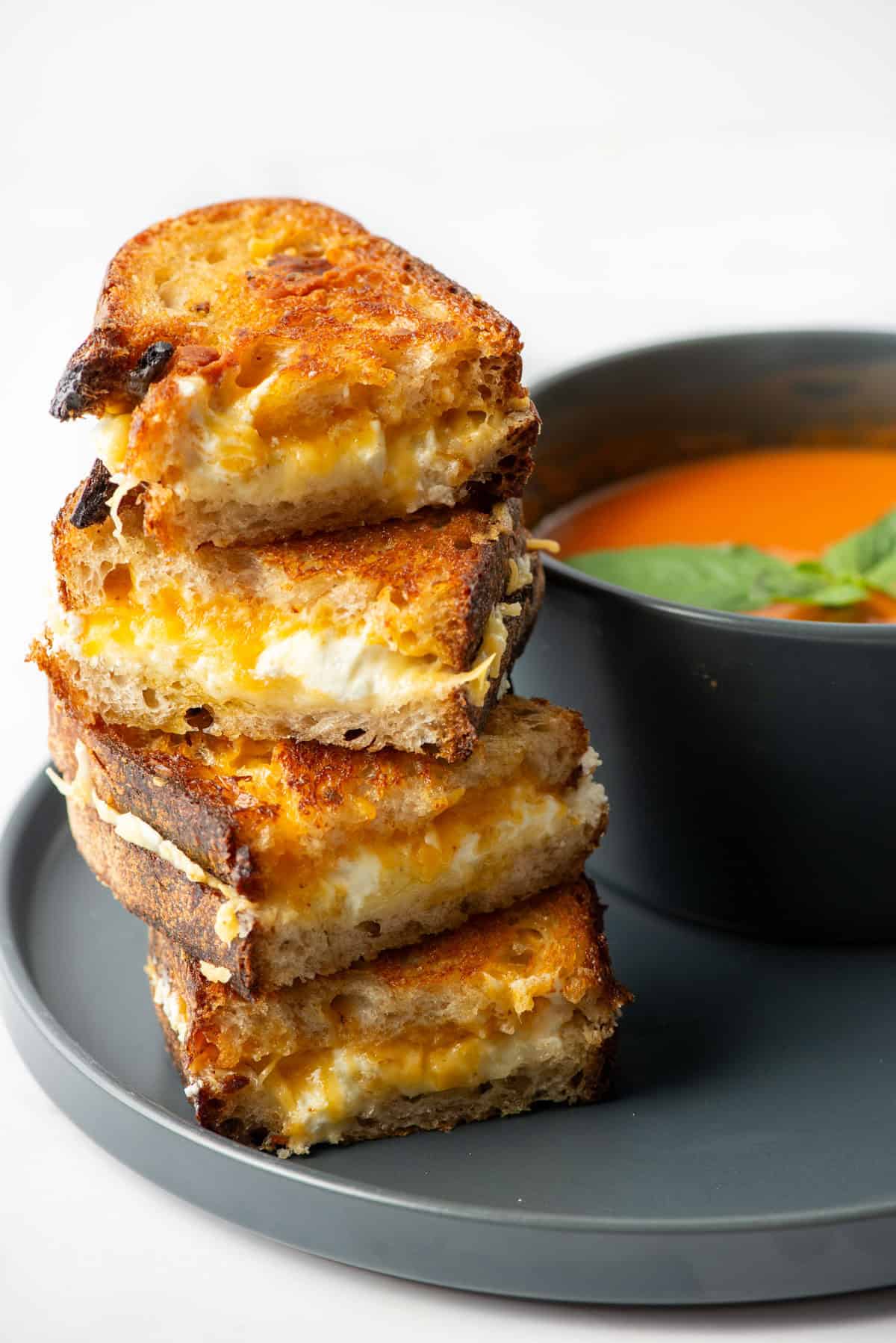 Side photo of a stack of four halves of grilled cheese sandwiches stack on top of each other next to a bowl of tomato soup