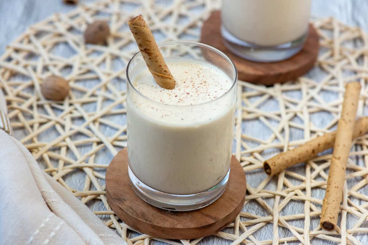 A frozen Brandy Alexander cocktail in a lowball glass with fresh grated nutmeg on top and a Pirouette cookie as a stir stick with another cocktail partially visible in the background