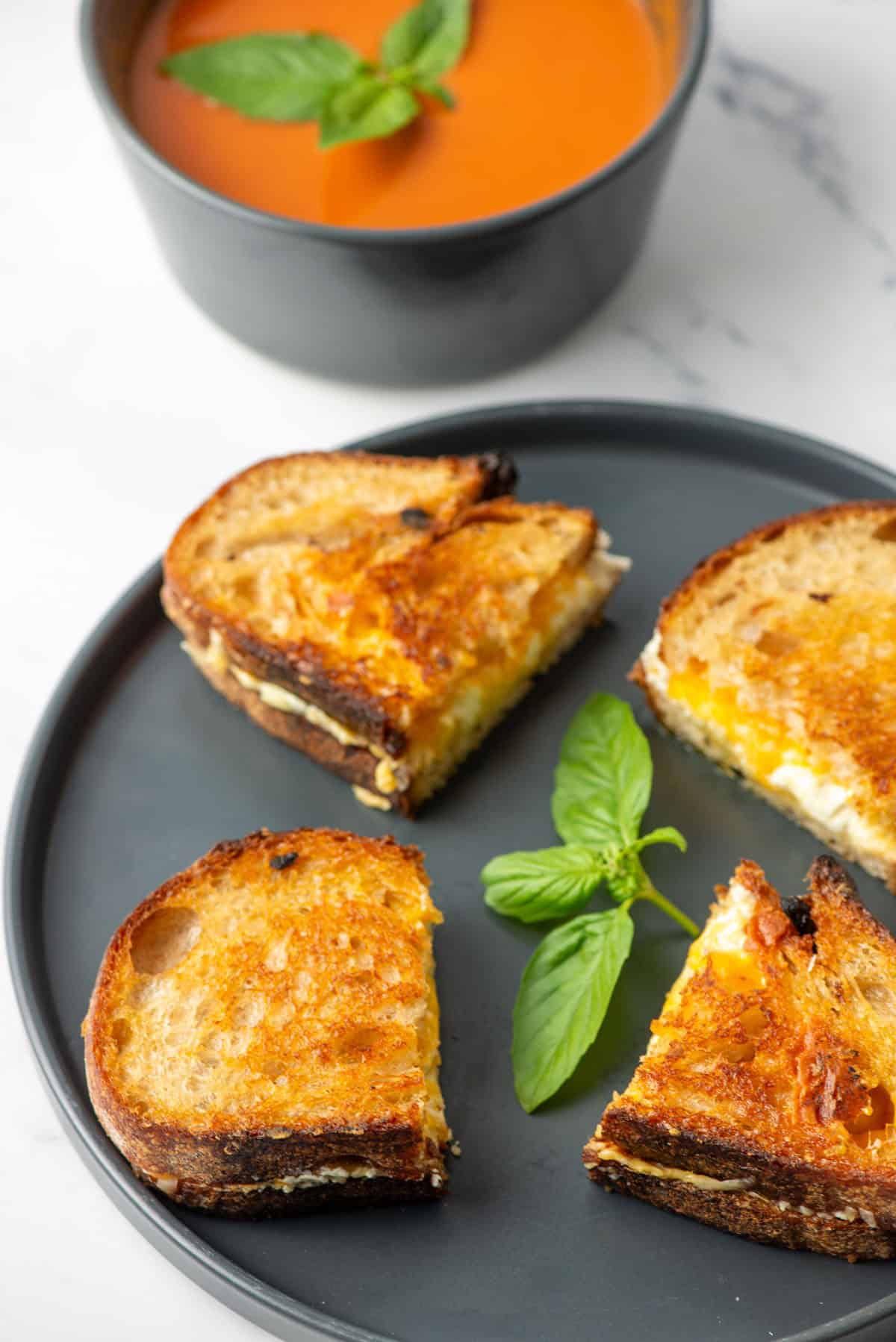 Two four cheese grilled cheese sandwiches on a gray plate in front of a bowl of tomato soup.