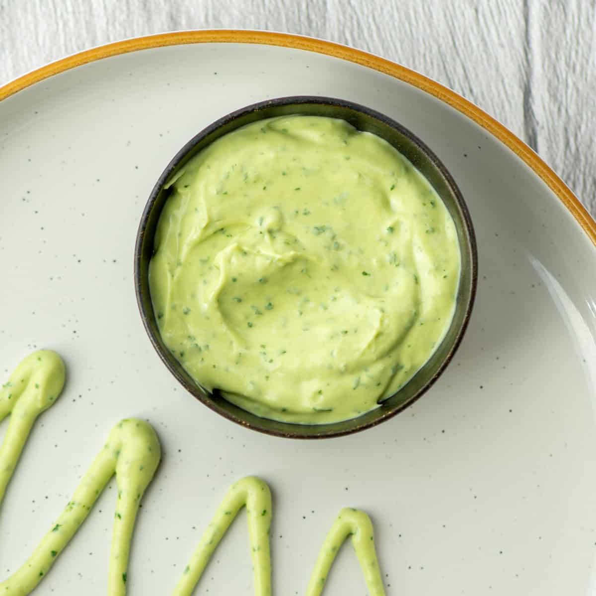 Overhead photo of a small sauce cup of avocado lime crema next to avocado lime crema drizzled on a plate in zig zag lines
