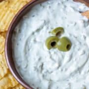 Bowl of dirty martini dip garnished with olives surrounded by kettle chips with a title overlay saying Dirty Martini Dip and another overlay at the bottom saying www.nextlevelbaker.com