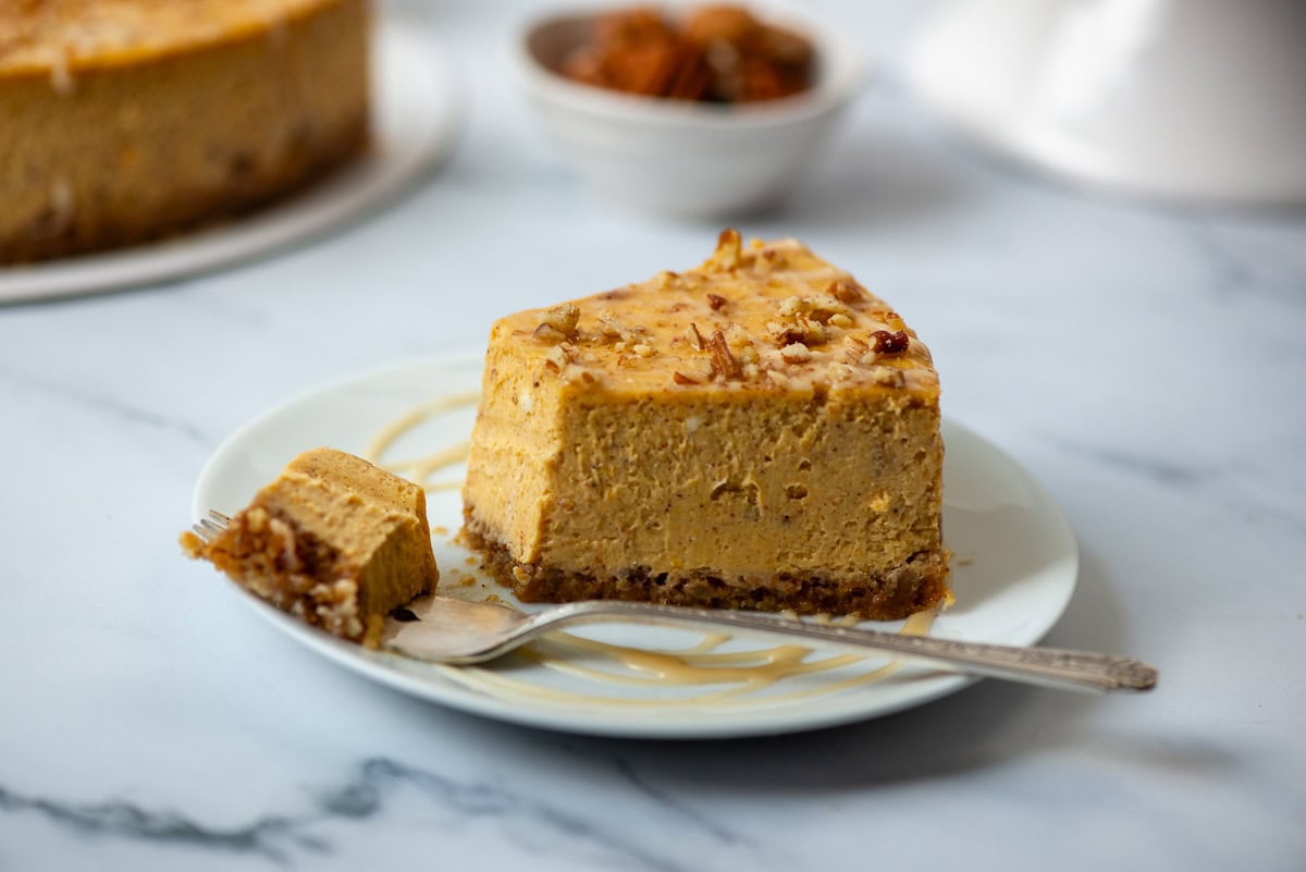 Side view of a slice of pumpkin pecan cheesecake on a plate in with a bite taken out and resting on a fork. A small bowl of almonds and the rest of the pumpkin pecan cheesecake is in the background
