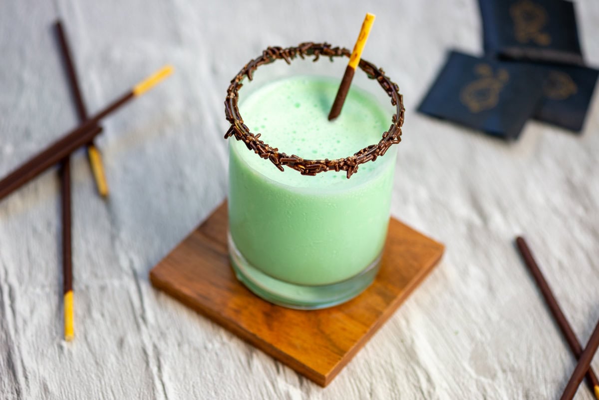 Close up photo of a frozen grasshopper drink with a chocolate sprinkle rim and a Pocky stick as a stir stick on a wood coaster with Pocky sticks and After Eight mints scattered in the background