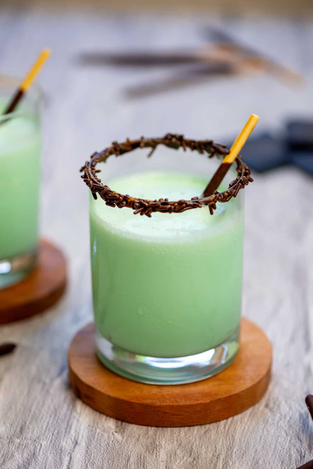 Sideview of a frozen grasshopper drink in a cocktail glass on a coaster with another frozen grasshopper drink and Pocky sticks in the background